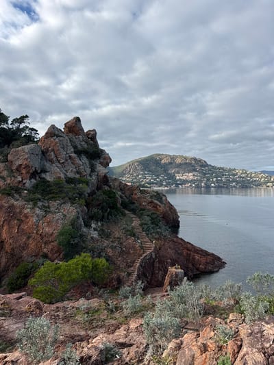 The image displays a coastal landscape in Théoule-sur-Mer, France, under an overcast sky. The foreground and midground are dominated by rugged, reddish-brown rock formations typical of the Esterel Massif, partially covered with sparse green and grey-green vegetation. A constructed stone staircase descends along the face of these rocks towards the calm, dark blue-grey waters of the Mediterranean Sea. Across the bay in the background, a large, green, tree-covered hill rises, densely populated with numerous white residential structures. No individuals are present. A small, dark, unidentifiable object is visible on the water's surface in the mid-distance.
