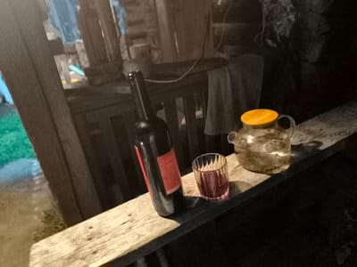 In Кашино, Russia, on a wooden railing, a dark glass bottle of red liquid stands next to a faceted glass filled with the same red liquid. To the right of the glass, a clear glass teapot with a bright orange lid sits. The background features rustic wooden structures and indistinct blurry elements. The scene appears to be outdoors, possibly in a rural or semi-outdoor setting.