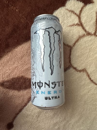 This is a close-up, overhead shot of a silver Monster Energy Ultra can lying on a fuzzy, textured surface that appears to be a blanket or rug. The surface has a pattern of light beige and brown shapes. The can is oriented horizontally, with its label facing the viewer. The Monster Energy logo, a stylized "M" in black and silver, is prominently displayed. To the left of the logo, in smaller text, there is writing in a foreign language, appearing to be Cyrillic script, that reads "БЕЗ ЦУКРУ" which translates to "SUGAR FREE". To the right of the logo, the words "MONSTER ENERGY" are visible, followed by "ULTRA" in a distinct font. The can itself has a silver base with intricate, light grey swirling patterns. The lighting suggests it is daytime, with shadows cast from the can onto the textured background. There are no people or other objects visible in the frame. The overall impression is of a casual, indoor setting. The location context provided is Cojușna, Moldova, which is a geographical identifier.