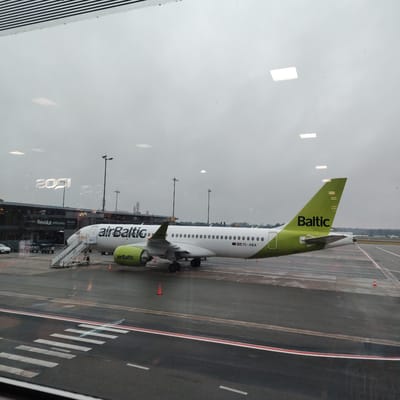 This image captures an AirBaltic Airbus A220-300 aircraft (registration YL-ABA) parked on an airport apron, viewed through a window from an indoor location. The scene is likely Riga International Airport, serving Skulte, Latvia, under an overcast sky.

The aircraft is predominantly white with prominent lime green accents on its tail fin, engine nacelles, and integrated into the "airBaltic" text on the forward fuselage. The word "Baltic" is also visible in lime green on the tail. A set of mobile boarding stairs, marked "L77", is positioned at the aircraft's front left door. The tarmac beneath the aircraft appears damp in areas and features white and red painted markings, including a pedestrian crossing with white stripes. Several orange traffic cones are placed on the apron.

In the background, a terminal building with "Revolut" branding is visible, along with multiple tall floodlight poles. Various ground service equipment and a white vehicle are partially visible near the terminal. Distant trees line the horizon under a muted, cloudy sky.

The window through which the image was taken exhibits significant reflections, including multiple square and rectangular interior lights, abstract patterns, and reversed text (e.g., "2091"). A horizontally striped pattern, possibly from blinds or architectural elements, is visible at the very top edge of the frame. No individuals are clearly discernible in the image.