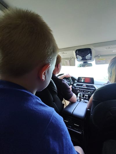 A group of people are inside a car on a road, possibly on a trip. The image is taken from the perspective of someone in the backseat. A young boy with blonde hair and wearing a blue shirt is in the foreground, his head and shoulder visible. To his right, a man with brown hair is driving, and a woman with blonde hair is in the passenger seat. The car's interior features a digital dashboard with a screen displaying a video, air conditioning vents, and a gear stick. The car appears to be moving through a rural or suburban landscape, possibly near Kurakino, Russia, with the bright daylight suggesting it's daytime.