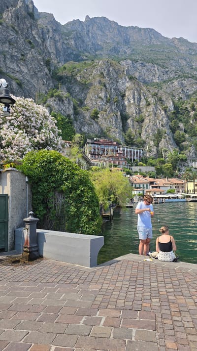 This media file shows a scenic view of a lake with a rocky mountain in the background, and a town with colorful buildings lining the shore. Two people are visible on a paved walkway next to the water. One person, wearing a light blue t-shirt and shorts, is holding something yellow and appears to be looking at it. The other person, wearing a black top with white polka dots and a skirt, is sitting down. The scene is bathed in sunlight, suggesting it's daytime. The lush green vegetation and blooming pink flowers add to the picturesque setting. The overall impression is one of a relaxed day in a beautiful Italian lakeside town, likely Limone sul Garda, given the context.