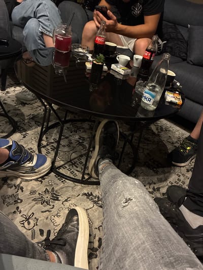 In a dimly lit, indoor setting, possibly a lounge or cafe, at the resort town of Tsigov Chark, Bulgaria, four individuals are casually seated around a circular, reflective black table. Their legs and feet are visible, suggesting a relaxed and informal gathering.
One person, positioned in the foreground, wears distressed grey jeans and black sneakers with white soles. Another person, to the left, is clad in denim jeans and blue and white athletic shoes. The third individual, further back and to the right, is dressed in dark pants and black sneakers. The fourth person's attire is less clear due to cropping.
On the table, several items are present: a glass mason jar filled with a dark red beverage and a black straw, two bottles of Coca-Cola, one open and one closed, a clear bottle of water with Cyrillic labeling, two white ceramic cups, an ashtray with cigarette butts, and two packs of cigarettes. A small potted plant adds a touch of greenery to the scene. The overall atmosphere appears to be one of leisure and social interaction, with a focus on beverages and smoking. The reflection on the table suggests a polished surface, possibly glass or high-gloss wood.