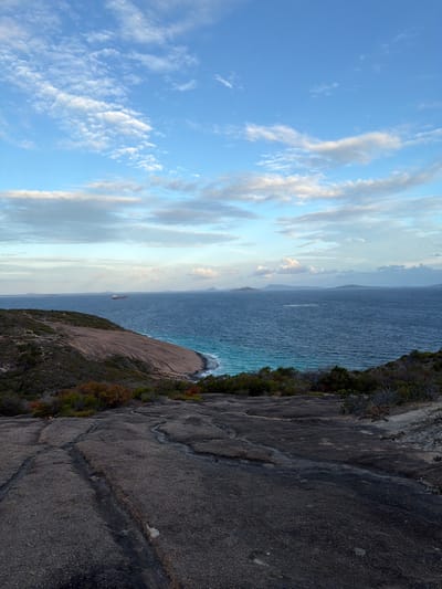 The image displays an expansive coastal landscape under a predominantly blue sky dotted with scattered white and grey clouds. In the immediate foreground, a dark grey, textured granite rock surface with visible natural cracks and undulations extends across the lower portion of the frame. Beyond this, a large, smooth, light brown granite outcrop descends towards the ocean, partially covered by sparse green and reddish-brown scrub vegetation. The deep blue ocean occupies the midground, transitioning to a bright turquoise hue closer to the shoreline where small waves are visible breaking. A prominent red and white cargo ship or tanker is present on the horizon in the left-midground, while several faint, dark landmasses or islands are visible along the distant horizon. The environment is rugged and natural, devoid of human structures or figures. The scene is located at West Beach, Australia.