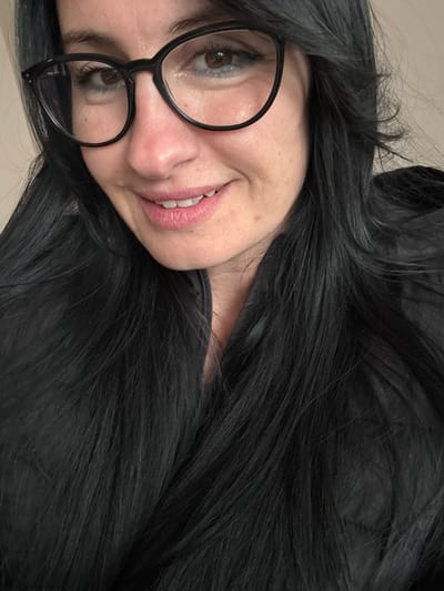A woman with fair skin and dark, shoulder-length hair is facing the camera with a slight smile. She is wearing black, round-framed glasses. She is wearing a dark jacket. The background is a light, neutral color. There is no indication of a location in Debrashtitsa, Bulgaria.