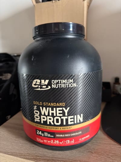 The image shows a large black tub of Optimum Nutrition Gold Standard 100% Whey Protein in Double Rich Chocolate flavor. The tub is prominently displayed on a wooden surface, possibly a shelf or table. The label on the tub is clearly visible, showing the brand logo, product name, and key nutritional information such as "24g HIGH PROTEIN PER SERVING" and "NET WT 2.26 kg e (5 LB)". The background is somewhat blurred but suggests an indoor environment with a light-colored wall and some out-of-focus objects. There is no indication of the city of Pravisdomini, Italy, or any people in the image. The image is a still photograph of a product.