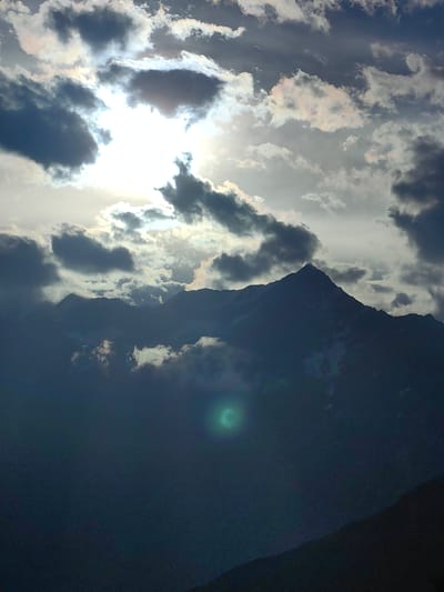 The image depicts a dramatic mountainous landscape under a cloudy sky. The sun is partially obscured by dark, ominous clouds, creating a high-contrast scene with bright light filtering through in places. Jagged mountain peaks are silhouetted against the sky, with some lighter, misty clouds clinging to their slopes. In the foreground, the lower slopes of the mountains fade into shadow, with a dark, indistinct foreground. There is a noticeable green lens flare in the mid-ground. The overall impression is one of a wild and powerful natural environment. The location is identified as Tiling, India.