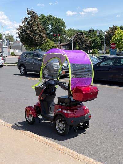 The image depicts a red mobility scooter parked on a paved surface, likely a street or parking lot, with a purple canopy featuring clear plastic windows and a bright yellow trim. The scooter has three wheels, a black seat, and a red storage box mounted on the back. It is positioned in the foreground, with other vehicles and trees visible in the background. A grey SUV is parked to the left, and a dark sedan is parked to the right. Traffic lights and a red "ARRÊT" (STOP) sign are visible further down the road, indicating an intersection. The sky is blue with scattered white clouds. Based on the "ARRÊT" sign, the location is Quebec, Canada. Given the specific visual elements such as the architectural style of the houses and the type of street infrastructure, this scene is likely from the city of Longueuil, Quebec, Canada. There are no people clearly visible on or near the scooter.