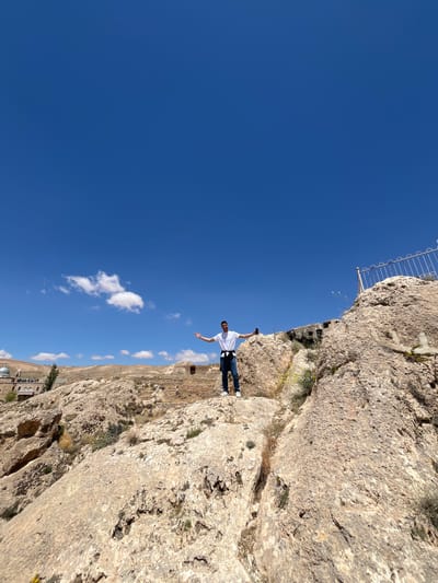 A man is standing on a rocky cliff with his arms outstretched, seemingly balancing himself. He is wearing a white shirt and blue jeans. The scene is oriented sideways. In the background, a blue sky contrasts with the beige-colored rock formations. The terrain below the cliff shows a valley or open area with some structures and sparse vegetation, hinting at a rugged landscape. A metal safety barrier is partially visible at the cliff's edge. The scene appears to be in a natural or semi-natural environment, with the possibility of a human-built structure visible in the landscape below. Given the information, it is plausible that this image was taken near Ma'loula, Syria, due to its mountainous terrain and historical sites.