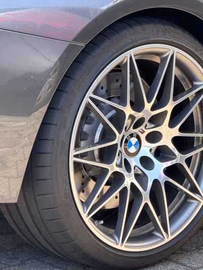 A close-up shot focuses on a BMW car wheel and tire. The silver rim has a complex, multi-spoked design with the BMW logo at the center. Perforated brake rotors are visible behind the rim. The black tire has a low profile and is partially visible. The car's gray body panel appears in the lower left corner. The tire's sidewall markings are partially visible.