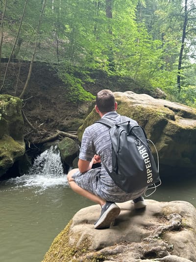 In the heart of Waldbillig, Luxembourg, a young man is captured in a moment of serene contemplation by a cascading waterfall. He is crouched on a moss-covered boulder, his back to the camera, facing the gentle rush of water. His attire, a patterned shirt and shorts, speaks of casual adventure, while a black backpack rests on his shoulders, emblazoned with the Superdry logo. The environment is a symphony of nature's artistry: lush green trees form a verdant canopy, framing the scene with an ethereal glow. Moss-laden rocks and gnarled roots create a natural stage for this tranquil tableau, with the waterfall serving as the focal point, its white spray a stark contrast against the earthy tones of the landscape. The water, a murky green, flows into a calm pool, reflecting the verdant surroundings. This is a moment of quiet communion with nature, a pause from the ordinary, set against the picturesque backdrop of Waldbillig's natural beauty.