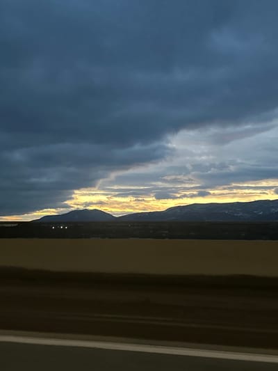 A twilight scene shows a range of dark, layered clouds taking up a majority of the view. A golden streak lights up the horizon where the clouds meet the mountains. The mountains are silhouetted against the bright sky. The bottom edge of the mountains shows a few faint lights, perhaps from a town or city. A portion of a window frame is on the right side of the image. The entire image is oriented vertically.