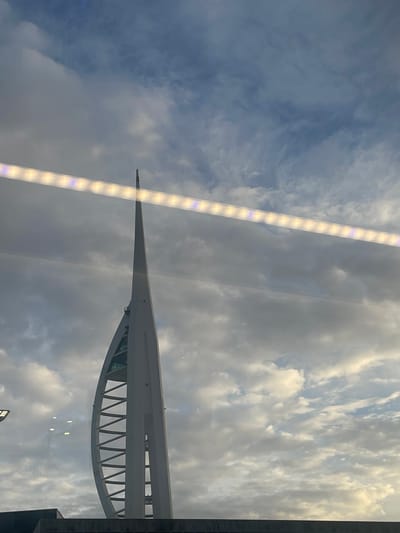 The image displays a prominent architectural landmark, the Spinnaker Tower in Portsmouth, United Kingdom, set against a cloudy sky. The tower is a tall, slender white structure with a distinctive pointed top and a broad, curved base featuring a geometric lattice design.

The sky is a dominant feature, filled with a mix of grey and white clouds, some appearing bright from sunlight while others are darker and more diffused. The overall impression is of a partly cloudy or overcast daytime scene, possibly late afternoon, given the quality of light.

The scene appears to be viewed through a reflective surface, likely a window. This is indicated by a bright, linear light fixture reflecting prominently across the left side of the frame. There are also other fainter, indistinct reflections visible, especially towards the bottom right. No people or specific activities are depicted. While some letters, possibly "TSW," are faintly visible on a horizontal bar of the tower's structure, they are not clearly legible due to the distance and angle. The image captures the modern architecture of the tower juxtaposed against the natural elements of the sky.