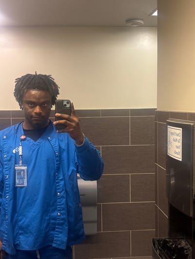 A person is taking a selfie in a bathroom. The person has dark hair and is wearing a blue button-down shirt, and a lanyard with an ID badge. The bathroom has light beige walls on the left side, and gray rectangular tiles with light grout on the right side. A paper towel dispenser is mounted on the wall, as well as a trash can. A toilet paper dispenser is visible in the background. Without further information, pinpointing the location of this bathroom in Grand Prairie is not possible.