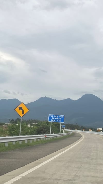 The image shows a highway in Baksukon, Indonesia. A gray asphalt road curves to the right, with a white lane marker dividing it. A gray metal guardrail runs along the edge of the road. On the left side of the road, two road signs are visible. One is yellow and diamond-shaped, with a black arrow curving to the left. The other is blue with white text in Indonesian, which translates to "Buses and Trucks Use Left Lane." In the background, mountains are visible under a cloudy sky. Green vegetation is seen between the road and the mountains.