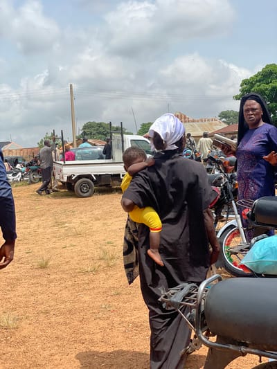 In Ban, Nigeria, a group of people are gathered around a small white pickup truck. A woman in a dark blue dress with intricate silver and pink patterns, wearing a black head covering, stands facing the viewer. She holds a silver object in her right hand and gestures with her left. To her left, a person in a black garment carries a young child on their back. The child wears yellow pants and has their arms around the carrier. Behind them, several motorcycles are parked. In the background, there are buildings with corrugated metal roofs and trees. The ground is covered in reddish-brown dirt. The sky is cloudy.