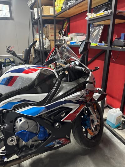 The image shows a BMW S 1000 RR motorcycle in a garage or workshop setting. The motorcycle is characterized by its white, red, and blue livery, carbon fiber accents, and high-performance components such as blue-colored engine covers and brake calipers. The background consists of shelving units containing various items, including boxes and containers. The location is likely a private garage or workshop, and there's no information to specify the city of Дубровка, Russia.