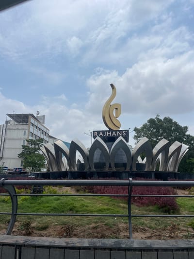 The image shows a golden sculpture with the word "RAJAHANS" written on a dark background in front of a building. The sculpture is located in the city of Ahmedabad, India. It is an outdoor scene with a cloudy sky. The sculpture is part of a larger structure that resembles a lotus flower with metallic petals. There is greenery and some bushes around the base of the sculpture. In the background, there is a multi-story building with windows and an antenna on the roof, as well as trees. A metal railing is visible in the foreground, separating the viewer from the landscaped area. No people are visible in the image.