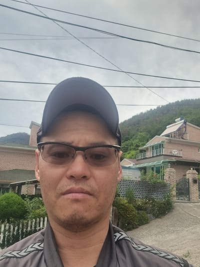 A man wearing glasses and a baseball cap takes a selfie outdoors. He is wearing a dark gray shirt with reflective chevron patterns on the shoulders. Behind him, a two-story house with a brick exterior and green-tinted windows is visible, situated on a hillside covered with green trees. The sky is overcast, and power lines crisscross above. The setting appears to be a residential area, possibly in Miryang-si, South Korea.