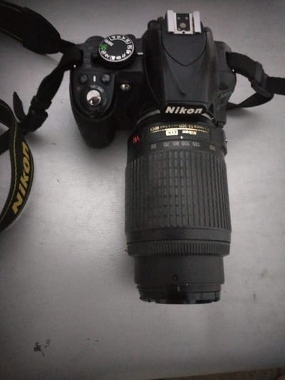 A black Nikon digital SLR camera with a telephoto lens is displayed against a light gray, textured surface. The camera is angled slightly, with the top and side visible. The Nikon logo is prominently displayed on the body of the camera and also on the lens barrel. The lens barrel has markings indicating focal lengths from 55mm to 200mm, as well as "VR" for Vibration Reduction. The camera strap, also black with yellow accents bearing the Nikon logo, is visible. A mode dial with various shooting mode icons is located on the top of the camera. The scene appears to be indoors or in a shaded area, given the lighting. There are no people in the image, and no specific time of day or weather conditions can be determined. The overall impression is that of a product shot, showcasing the camera equipment. The visible text includes "Nikon," "NIKKOR 55-200," "VR," "DX," and various shooting mode labels on the dial such as "GUIDE," "M," "A," "S," and "P."