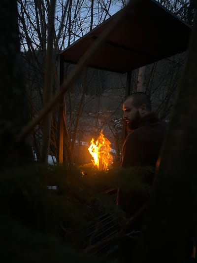 In an outdoor setting in Korostiv, Ukraine, captured at dusk or night, a male individual with a beard and short dark hair is depicted in profile, facing right. He wears dark clothing and stands to the right of a large, brightly burning fire. The fire, characterized by intense orange and yellow flames, is contained within a dark metal structure topped with a square canopy, under which the man is partially positioned. The firelight illuminates the man's face and the immediate surroundings. Behind the man and the fire, a natural environment is visible, featuring numerous bare deciduous trees silhouetted against a pale blue-grey twilight sky. Patches of snow are present on the ground, and a darker, dense forest or hillside recedes into the distance. The immediate foreground is partially obscured by dark, out-of-focus tree branches and needles.