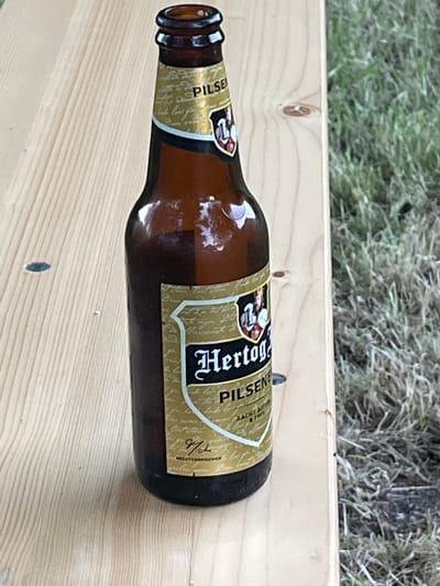 A close-up, eye-level shot shows a brown glass bottle of Hertog Jan Pilsener beer resting on a light-colored wooden surface. The bottle is positioned vertically, with its neck pointing upwards and slightly to the left. The label on the bottle is predominantly gold with black and red accents. The word "PILSNER" is visible in large black lettering on the upper part of the label. Below this, a crest-like emblem with figures is present, followed by the name "Hertog Jan" in stylized black font. The wooden surface appears to be a tabletop or bench, with visible wood grain and a few small dark specks and circular indentations. To the right of the bottle, the background consists of out-of-focus green grass and some darker, drier patches of ground, suggesting an outdoor setting. The lighting indicates it is daytime. There are no people visible in the image. The scene does not provide any specific contextual information that would allow for identification of the location in Uithuizen, Netherlands.