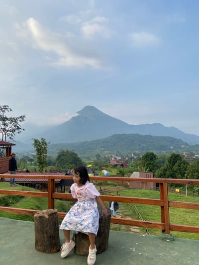 A girl wearing a light pink top and a light blue floral dress is leaning over a railing made of wooden planks and sitting on a pair of tree stumps. The railing is positioned on an elevated platform with a green surface. In the background, a mountainous landscape is visible, with trees, buildings, and tents dotted across the terrain. The sky is a light blue color with some clouds. The location appears to be in or near Tamiajeng, Indonesia.