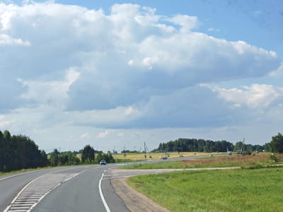 The image captures a rural highway scene near Podcherkovo, Russia, under a partly cloudy sky. A paved road curves from the foreground into the middle distance. White dashed lines mark the lanes and solid white lines delineate the edge of the road. A section of the road features a wide, striped pedestrian crossing. To the left of the road, a dense line of dark green trees forms a natural barrier, while to the right, a bright green grassy field extends outwards. In the background, a broad, yellowing field stretches towards a distant line of trees. Several vehicles are visible on the road, including a dark sedan in the left lane, a white car further ahead on the right, and what appears to be a truck and another vehicle in the distance. Utility poles with wires are scattered throughout the landscape. The overall impression is one of a serene, sunny day in the Russian countryside.