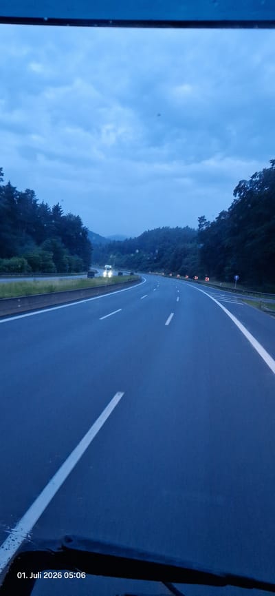 A highway stretches through a wooded, hilly landscape under a cloudy, twilight sky. The road is wet, indicating recent rain, with visible white lane markings and a solid white line marking the shoulder. On the left side, a grassy embankment borders the highway, backed by dense evergreen trees. On the right side, the highway curves, with a barrier and traffic signs visible. A single truck with illuminated headlights is approaching in the distance on the opposite side of the highway. The image appears to be taken from a moving vehicle, with the bottom edge showing a portion of the vehicle's dashboard or windshield. A timestamp in the lower left corner reads "01. Juli 2026 05:06". The location is identified as Betzenstein, Germany.
