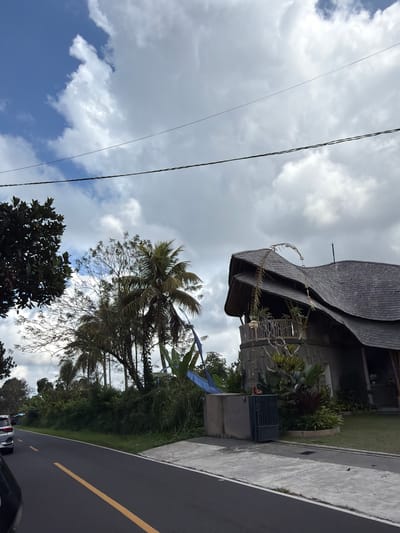 In Susut, Indonesia, on a cloudy day, a rural road curves gently to the left. The road is paved with asphalt and marked with a single yellow line. On the right side of the road, a unique, modern house with a thatched roof and a curved design stands prominently. The house has an open balcony with plants and decorative elements. In front of the house, there is a grey metal gate and a concrete structure. Lush green vegetation, including palm trees and various plants, lines the roadside. A silver SUV is visible on the road, indicating movement. Overhead, power lines stretch across the frame against a backdrop of scattered clouds in a blue sky. The scene captures a blend of natural tropical scenery and contemporary architecture.