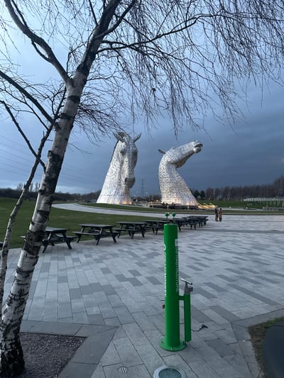 The image depicts an outdoor scene at dusk or early evening, located in Helix Park, Falkirk, United Kingdom, dominated by two large, illuminated, silvery metallic horse head sculptures known as "The Kelpies."
In the immediate foreground, to the left, is the trunk and bare branches of a birch tree. The ground consists of a paved area featuring light and dark grey rectangular pavers, upon which are arranged multiple dark-colored wooden picnic tables with attached benches, all currently unoccupied. To the right of the paved area stands a bright green cylindrical public bicycle repair station, marked "GREASEMONKEY" and "INFRASTRUCTURE" vertically, equipped with a bicycle pump and tools. A small circular ground light is visible near its base.
Behind the paved area and picnic tables, a manicured green lawn extends towards the sculptures. A curved paved pathway or road traverses this grassy expanse.
The midground is occupied by the two monumental horse head sculptures. The sculpture on the left faces primarily forward, angled slightly to the right. The sculpture on the right features a raised head with a slightly open mouth, angled towards the right. Both sculptures are illuminated from their bases with warm white light.
In the background, beyond the sculptures, the landscape includes scattered leafless trees and visible utility poles with power lines on the far left. To the far right, a small group of three indistinct individuals is observed standing on a paved area, oriented towards the sculptures. The sky above is dark and overcast, suggesting an approaching night.