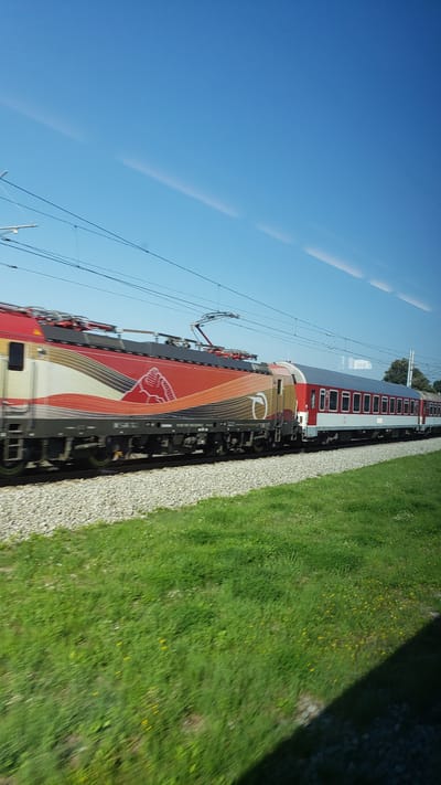 This is a side view, captured from a moving train, of a red and white passenger train traveling on a track. The train has a distinctive red locomotive with a stylized white and orange design that includes a depiction of mountains. It is connected to several red and white passenger cars. The train is traveling through a rural landscape with a clear blue sky above. In the foreground, there is a strip of green grass and a gravel track bed. The image appears to be taken on a sunny day. The location is stated to be the city of Varín, Slovakia.