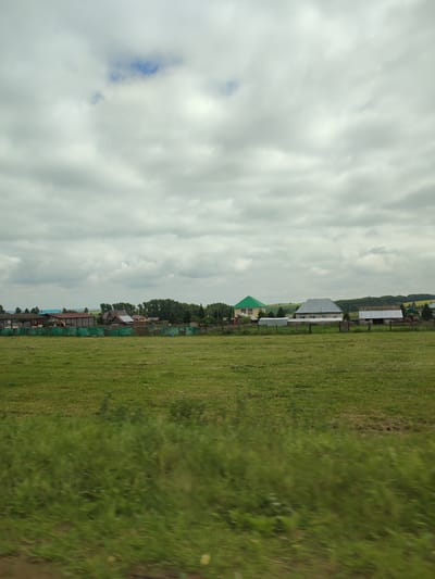 The image depicts a rural landscape with a village in the background and a lush green field in the foreground. Several houses with different colored roofs, some with garages, are visible, suggesting a residential area. A green fence runs along the edge of the village, separating it from the field. Trees and bushes form a backdrop to the houses, and in the distance, rolling hills are visible. The sky is overcast with scattered clouds, indicating a cloudy day. The overall scene conveys a sense of tranquility and rural life. There is no visible text in the image. The location is Verkhneyarkeyevo, Russia.