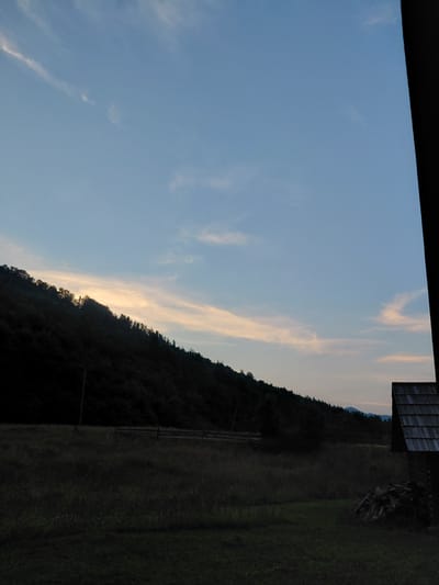 The image captures a serene twilight scene in Chernyk, Ukraine. The sky is a gradient of soft blue, transitioning to a pale orange where the sun has recently set, with wispy clouds streaking across the horizon. A dark, tree-covered hillside dominates the mid-ground, its silhouette stark against the fading light. To the right, a field of green grass, likely a meadow or pasture, stretches out, appearing lush even in the dimness. At the top right of the frame, the corner of a wooden structure, possibly a barn or shed, is visible, adding a touch of human presence to the otherwise natural landscape. The overall mood is peaceful and tranquil, evoking the quiet beauty of a rural evening. There is no visible text in the image.