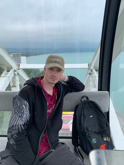 A man is reclined in a seat inside a Ferris wheel carriage. He is wearing a dark-colored hooded sweatshirt with a wing design on the sleeves, a red collared shirt underneath, and a green baseball cap. A black Puma backpack is next to him. Outside the window, a body of water and some buildings are visible. Based on the image's context, it is likely that this image was taken on the Ferris wheel in Bichvinta, Abkhazia.