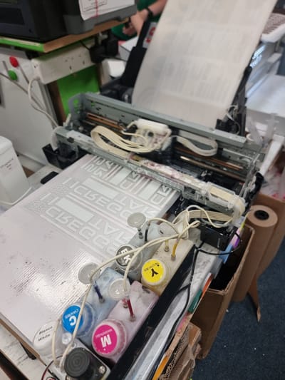 This image depicts a scene inside a printing workshop, likely a small business, possibly located in or near Tullamore, Ireland. The foreground is dominated by a printer and associated equipment. A white sheet of paper or substrate is laid flat on a surface, with several white graphic elements printed on it, possibly for signage or custom decals. To the right of the paper, a row of ink bottles is visible, containing colored inks labeled in English and possibly another language. These include Cyan, Magenta, and Yellow. The bottles are connected by tubes to the printer mechanism.
The printer itself appears to be a large format inkjet printer, with its print head mechanism exposed. Ribbons and wires are visible, suggesting it's in operation or has recently been used. Behind the printer, a roll of brown paper or similar material is standing upright in a cardboard box. The background shows a cluttered workshop environment with shelves, electrical outlets, and other printing-related machinery. A person's arm, wearing a dark sleeve, is partially visible in the upper right corner, suggesting someone is present and potentially working in the vicinity. The overall impression is of an active, if somewhat disorganized, workspace dedicated to printing.