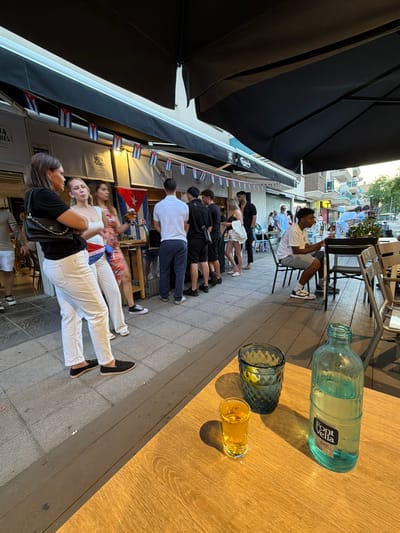 The image depicts an outdoor setting in Cambrils, Spain. A group of people are seated along a sidewalk next to a building. Cuban flags are visible along the building's awning. A man is seated at a table, holding a phone. On the table, there is a water bottle, a glass with ice, and a shot glass with a yellow liquor.