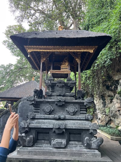 In the city of Dawan, Indonesia, a person's hands are visible in the lower left quadrant of the image, holding a mobile phone vertically to capture a photograph. They are wearing a blue long-sleeved shirt. The foreground features an intricately carved stone structure, possibly a temple or shrine, with multiple tiers and decorative elements, including what appear to be guardian statues shaped like winged beasts. Behind this structure, a multi-tiered roof with thatched covering and ornate golden detailing shelters an inner sanctuary. The background reveals lush green foliage and a rocky incline. To the left of the main structure, another thatched roof building is partially visible. The overall scene depicts a cultural or religious site bathed in natural light, with an overcast sky overhead.