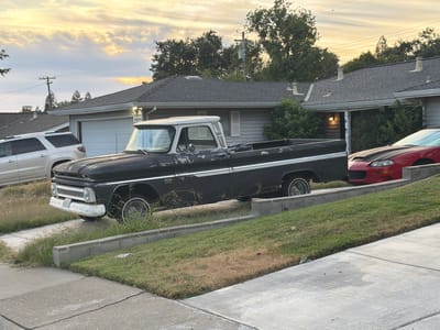 A black vintage pickup truck with a white roof is parked in a driveway. The truck is a Chevrolet C10 from the 1960s and features chrome accents and wire-spoke wheels. A white SUV is parked to the left of the truck, and a red sports car is to the right. The vehicles are parked in front of two houses with gray siding and dark gray roofs. The scene is a residential neighborhood, likely in Sacramento, California, given the context.

The time of day is late afternoon or early evening, with the sky displaying soft hues of orange and yellow from the setting sun, indicating a partly cloudy and pleasant weather condition. The sun's warm light illuminates the scene, casting soft shadows. The grass in the yard is somewhat dry and patchy, suggesting a warm climate. The overall atmosphere is peaceful and suburban.

There is no visible text in the image. The focus is on the vehicles and the residential setting.