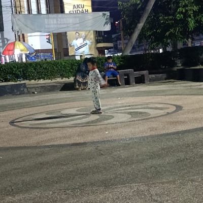 A young boy in pajamas stands on a circular, cobblestone paved area, looking towards the right. He is in the center of the image, with the camera slightly tilted downwards. The scene is lit by streetlights at night. Behind the boy, a person in a long garment is walking out of view to the left. Another person in a blue shirt and jeans is sitting on a bench to the right, looking down at a phone. The boy is the primary subject of the scene, with the other individuals in the background. There is an urban environment surrounding the scene. The location is a small park or square in Kecamatan Asembagus, Indonesia, at coordinates -7.75 latitude and 114.218 longitude. The image is captured from a low angle, and the lighting is soft, emphasizing the subject's silhouette against the background. There is a street sign with Indonesian text in the background. There are bushes and trees in the background. The scene appears to be peaceful and serene, with no signs of commotion or activity. The boy's posture and the overall calmness of the scene create a sense of quiet contemplation.