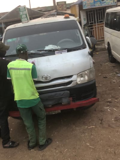 The image portrays an outdoor urban scene in the city of Tukur Tukur, Nigeria. The central subject is a white Toyota Hiace-style commercial van, viewed from the front, parked on reddish-brown dirt ground with scattered debris.
The van features a black front bumper with a prominent red-painted aftermarket bull bar or protective frame affixed below it. An orange rotating beacon light is mounted on its roof. A rectangular sign attached to the roof rack displays partially legible text including "NORTH PHONES LINE BOOK ACHA NIGERIA." The vehicle's windshield wipers are in the upright position, and a white cloth along with a stack of papers or tickets rests on the dashboard. The van shows signs of use and dirt.
Two men stand in the foreground, facing the front of the Toyota van. One man, viewed from the back, wears a bright lime-green reflective vest over a green t-shirt, green cargo pants, and black sandals. He wears a green baseball cap with the acronym "KURTW" and "KADUNA STATE" visible on the back. The second man, partially visible on the left, wears a dark shirt and black sandals.
In the background, commercial structures with corrugated iron roofs and various storefronts are present. One storefront displays metal security bars and indistinct signage, including a partially visible "DJ" graphic. To the right of the main van, a portion of another white van is visible. The sky appears overcast or hazy.
