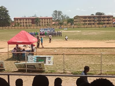 The image depicts an outdoor scene, likely a sports field in Isasi, Nigeria. A track surrounds a grassy field. In the foreground, a fence with a few signs and a tent are visible, with several people near the tent. On the field, two teams of athletes stand facing each other, with a banner or display board behind them. In the background, low buildings stand, suggesting a residential or institutional setting. The sky is bright, suggesting daytime. People appear to be watching from the foreground.
