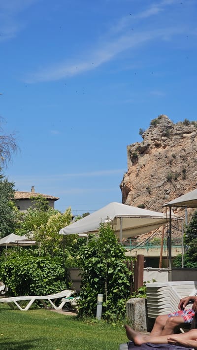 The image depicts an outdoor scene at what appears to be a resort or recreational area in Alhama de Aragón, Spain. In the foreground, a person's legs are visible, clad in plaid shorts, resting on lounge furniture. A neatly mowed green lawn occupies the lower portion of the frame, with a white plastic sun lounger positioned on it. Lush green bushes and trees are prominent throughout the mid-ground, interspersed with white canopies providing shade. To the left, a building with a tiled roof and a visible chimney can be seen behind the foliage. Dominating the background is a large, rocky cliff face with signs of past construction or stabilization. The sky is a clear, bright blue, dotted with numerous small, dark shapes that appear to be birds in flight.