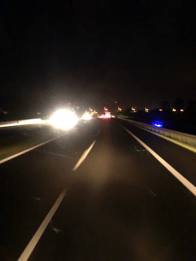 The image depicts a night-time scene on a highway, likely in or near Imielno, Poland, characterized by the blurred motion of vehicles and road markings. The dominant feature is a bright, overexposed glare of headlights from an oncoming vehicle, obscuring details of that specific car. Lane markings, a white line on the left and another on the right, are visible, indicating a multi-lane road. The surrounding environment is dark, with faint, blurry lights in the distance suggesting other vehicles and possibly buildings or streetlights. A bright blue light is visible on the right side of the road, near the barrier, its source and purpose unclear. The overall impression is one of speed and limited visibility due to the darkness and motion blur. There are no discernible individuals or specific objects clearly identifiable beyond the general context of a highway at night.