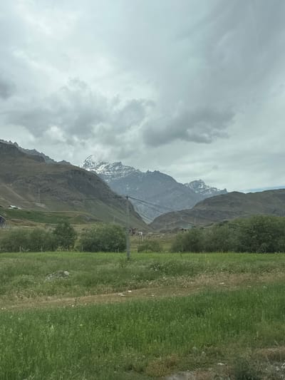 The image depicts a mountainous landscape in Dras, India, under a cloudy sky. The foreground features a lush green field, with a dirt path winding through it. Beyond the field, a line of trees and shrubs is visible, with some scattered structures, possibly houses or buildings, nestled on the green hillsides. In the midground, a utility pole with wires stretches across the scene, connecting to other poles. The background is dominated by imposing mountains, some of which are capped with snow. The overall atmosphere is serene and natural, characteristic of a rural mountainous region. There are no people or animals visible in the frame.