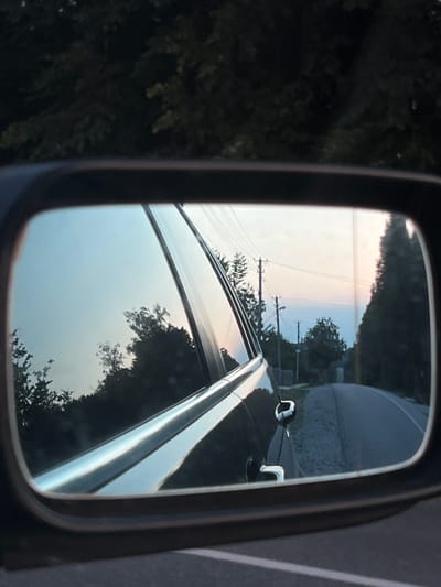 The image is a reflection in a car's side-view mirror, showing a road curving through a rural landscape. The sky is a gradient of pale blue to pink, suggesting either dawn or dusk. Trees and utility poles line the road. The reflection also shows a portion of the car's body, including a door handle. The context provided indicates this scene is located in Mshanets, Ukraine.