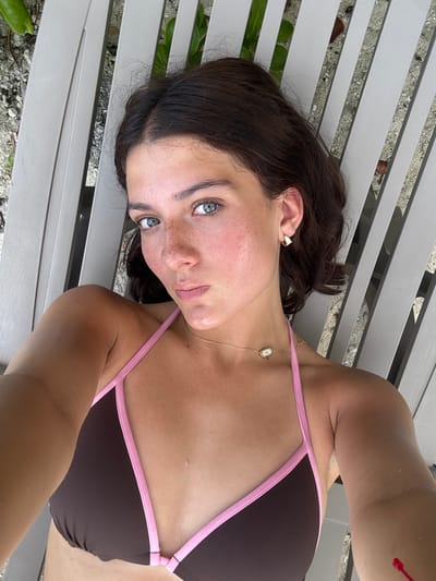 A young woman lies on a light gray bench, possibly made of wood or metal, outdoors. She is wearing a brown bikini with pink straps. She has brown hair, fair skin with freckles, and striking blue-green eyes. She wears a gold necklace with a pendant and small gold earrings. The background suggests an outdoor setting with greenery and possibly gravel or sand. The location is specified as San Miguel de Cozumel, Mexico. The woman is lying down and looking directly at the camera, suggesting a selfie.