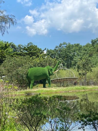 An artificial, green, elephant-shaped sculpture stands on a grassy bank next to a tranquil body of water. The sculpture, covered in what appears to be artificial turf, is positioned to the left of a large, wire-frame structure that resembles a basket or a bird's nest. This structure is also situated on the grassy bank. Behind the elephant and the wire structure, a dense line of lush green trees forms a backdrop. The sky above is blue with scattered white clouds. The water in the foreground reflects the trees and the sky, creating a mirrored image of the environment. The scene is set in Hoa Thang Ward, Vietnam.