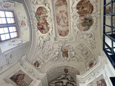 The image shows the richly decorated interior ceiling of a building in Náchod, Czechia. The ceiling features extensive white stucco ornamentation and several painted frescoes depicting religious or mythological scenes. The stucco work includes intricate scrolls, cherubs, and figures that appear to be angels or mythological beings. The frescoes are oval or rectangular in shape and are framed by the stucco. One prominent fresco in the upper center depicts a scene with multiple figures and cherubs, possibly a heavenly gathering. To the left, a large window with multiple panes allows natural light to illuminate the space, with smaller oval frescoes on the surrounding stucco. The perspective is looking upwards, emphasizing the grandeur and artistry of the ceiling. A dark metal railing or structure is visible on the right side of the image, suggesting a balcony or upper level overlooking the space. The overall impression is one of opulence and detailed craftsmanship.
