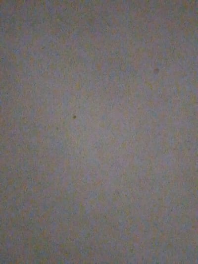The image is an abstract, low-resolution, noisy photograph of a textured surface. The dominant color is a muted gray with subtle hints of brown, yellow, and blue. There is a small, dark, circular spot visible slightly left of the center. The texture appears granular or speckled. The image does not contain any discernible features that would allow for identification of its location in Verkhniy Mamon, Russia, nor does it depict any people, subjects, objects, actions, or interactions. The environment is not discernible.